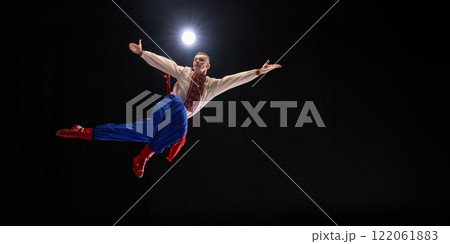 Performer leaping with arms upward, leg raised in dynamic move, traditional embroidered outfit expressing artistry and tradition against black studio background. 122061883