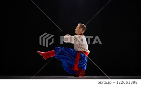 Male dancer in deep squat with one leg extended, arms elegantly balanced, showcasing agility and tradition against black studio background. 122061898