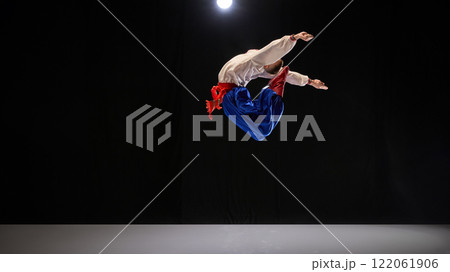 Male dancer executing backward jump, dressed in traditional embroidered shirt, blue trousers, and red boots, expressing culture and movement against black studio background. Male dancer executing backward jump, dressed in traditional embroidered shirt, blue trousers, and red boots, expressing culture and movement against black studio background. 122061906