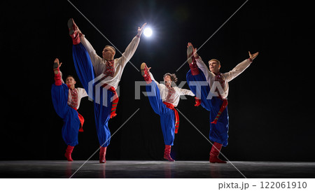Male dancers in powerful high kick pose, wearing embroidered shirts, blue trousers, and red boots, showcasing tradition and precision against black studio background. 122061910
