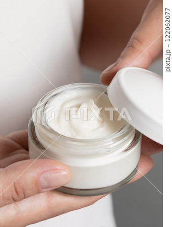 Woman opening cream jar against white shirt and grey background closeup 122062077