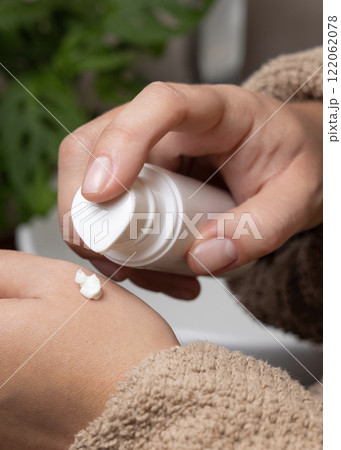 Woman in brown bathrobe applying cream on hand and holding a cosmetic bottle closeup 122062078