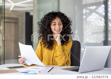 Satisfied businesswoman smiling holding report in hands, office worker among papers and contracts working inside office with laptop. Financier accountant with successful achievement results. 122062579