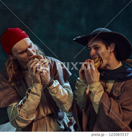 Close-up portrait of tow men, medieval pirates in hat and bandana, passionately eating burgers against dark vintage background Close-up portrait of tow men, medieval pirates in hat and bandana, passionately eating burgers against dark vintage background 122063537