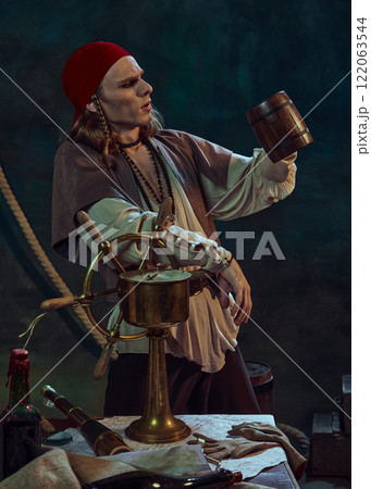 Young man, pirate in red bandana leaning on ship wheel, gazing into wooden mug with drink against dark background with maritime tools Young man, pirate in red bandana leaning on ship wheel, gazing into wooden mug with drink against dark background with maritime tools 122063544