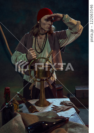 Young man, pirate in red bandana holding ship wheel, gazing into horizon with determined look against dark background with maritime tools Young man, pirate in red bandana holding ship wheel, gazing into horizon with determined look against dark background with maritime tools 122063546
