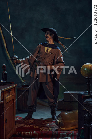 Lively pirate captain laughing confidently while looking at rapier against dark background with vintage maritime props. 122063572