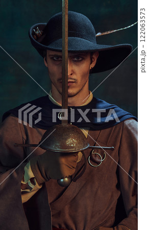 Portrait of handsome young man, pirate with dark eyes and intense gaze, in vintage costume and dark hat, holding sword next to his face 122063573