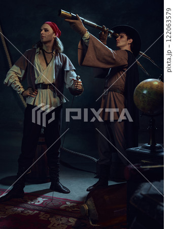 Brave pirates. Two men, dressed in vintage costumes, standing in a dark-lit rood with vintage items. One man stands with sword, while his companion looking in telescope. 122063579