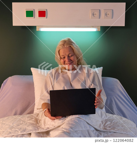 Happy smiling elderly woman sitting in hospital bed and chatting with family during online video call via laptop in the evening against green wall 122064082