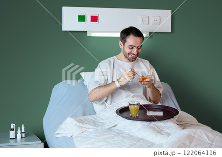 Male patient in white hospital gown sitting in bed with food tray, smiling, eating and drinking juice against green wall background. Male patient in white hospital gown sitting in bed with food tray, smiling, eating and drinking juice against green wall background. 122064116