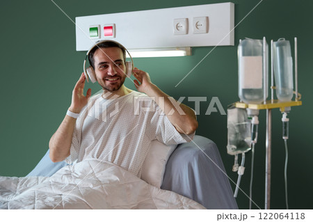 Male patient sitting in bed in hospital ward, wearing headphones and smiling, with IV drip against green wall with special emergency buttons Male patient sitting in bed in hospital ward, wearing headphones and smiling, with IV drip against green wall with special emergency buttons 122064118