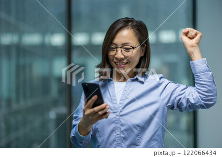 Successful Asian woman celebrating success and victorious achievement results. Businesswoman standing by window inside office building with phone in hands, received online notification of triumph. 122066434