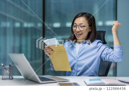 Joyful and successful Asian woman at workplace received mail notification letter envelope with good winning news, businesswoman sitting with laptop, holding hand up reading message. 122066436