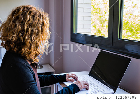 Blond curly hair adult woman working on her computer in front of the window. A coffee break Blond curly hair adult woman working on her computer in front of the window. A coffee break 122067508
