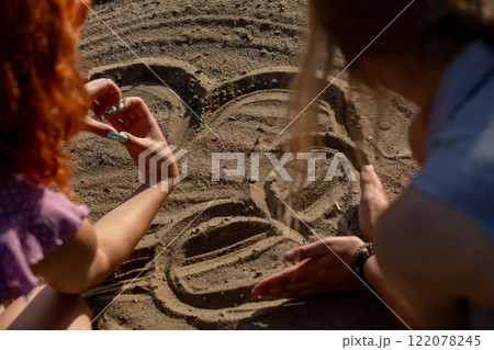 Two close friends happily drawing in the warm sand on a sunny beach, enjoying their time 122078245