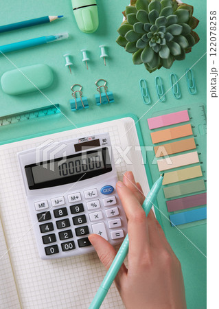 Organized workstation with office supplies in mint green theme. A woman's hand is pressing buttons on the calculator. 122078258