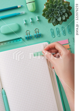 Hand sticks a post-it note in an open notebook with a blank page. Mint green stationery, pastel background. Hand sticks a post-it note in an open notebook with a blank page. Mint green stationery, pastel background. 122078259