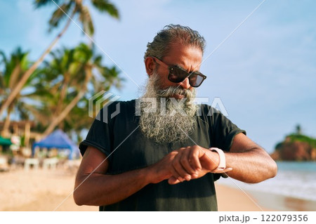 Stylish gray-bearded senior in sunglasses uses smart watch on tropical beach. Modern mature man explores tech gadgets. Active fitness lifestyle, relaxed vacation scene. Stylish gray-bearded senior in sunglasses uses smart watch on tropical beach. Modern mature man explores tech gadgets. Active fitness lifestyle, relaxed vacation scene. 122079356