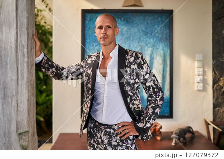 Bald man in floral suit leans on wall inside villa. Daytime setting with modern decor. Wedding attire. Calm expression suggests confident mood. Stylish groom or fashion statement theme. Bald man in floral suit leans on wall inside villa. Daytime setting with modern decor. Wedding attire. Calm expression suggests confident mood. Stylish groom or fashion statement theme. 122079372