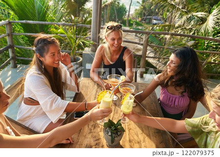 Group of diverse women friends toast drinks on sunny terrace with ocean view. Female brunch gathering. Girls club meeting. Socializing outdoors with cocktails. Relaxing in tropical restaurant. 122079375
