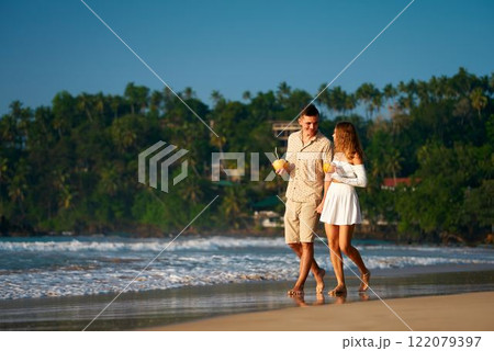 Romantic couple walk on sandy beach, man and woman hold tropical drinks, stroll by sea during sunset. Barefoot honeymooners enjoy ocean waves, relaxed vacation. Love, travel, summer concept captured. 122079397