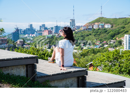 A 50-year-old Russian woman in a white dress sits on wooden steps in Nagorny Park, Vladivostok, enjoying the cityscape and greenery under the summer sun 122080483