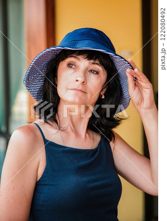 A 50-year-old Russian brunette woman wears a blue sun hat and adjusts it while looking to the side. Close-up portrait showing summer style, wrinkles, and elegance in warm weather A 50-year-old Russian brunette woman wears a blue sun hat and adjusts it while looking to the side. Close-up portrait showing summer style, wrinkles, and elegance in warm weather 122080492