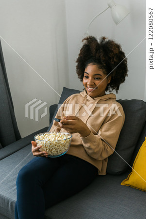 Young african american woman with a remote control in one hand and popcorn watches a television program. Cheerful enjoyment of leisure time. Streaming service, film and tv show concept. Young african american woman with a remote control in one hand and popcorn watches a television program. Cheerful enjoyment of leisure time. Streaming service, film and tv show concept. 122080567