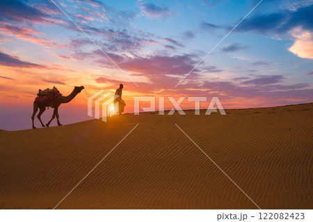 Indian cameleer camel driver camel silhouettes in dunes on sunset. Jaisalmer, Rajasthan, India Indian cameleer camel driver camel silhouettes in dunes on sunset. Jaisalmer, Rajasthan, India 122082423
