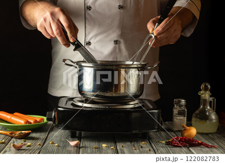 An experienced cook expertly maneuvers a knife and fork while stirring the contents of a pot on the stove. Fresh ingredients such as onions and garlic are nearby, adding color to the cooking space 122082783