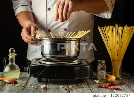 An experienced cook sprinkles salt over pasta in a boiling pot, surrounding it with fresh ingredients such as onions and herbs, demonstrating culinary skill during a cooking session 122082784