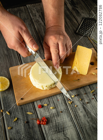 Skilled hands slice a round cheese on a wooden board with fresh lemon and spices scattered around. The warm kitchen exudes a cozy atmosphere, perfect for culinary creation 122082786