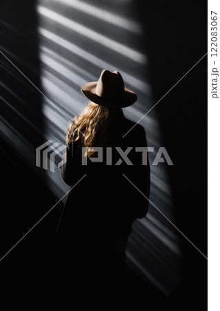 A lone blonde woman in the dark wearing a coat and a hat, seen from behind. A dark figure of an incognito person standing in the rays of light. AI-generated 122083067