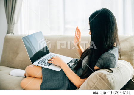Asian woman on sofa uses laptop for video conferencing saying hi. Engaged in distant meeting discussing and smiling. Modern communication and connection online. Asian woman on sofa uses laptop for video conferencing saying hi. Engaged in distant meeting discussing and smiling. Modern communication and connection online. 122083711