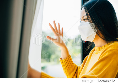 Depicting COVID-19 isolation, Woman in a protective mask gazes through the window feeling upset and lonely. Reflecting fear solitude and the effects of the pandemic crisis. 122083712