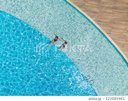 Two kids relax at the edge of clear blue pool during sunny summer vacation. This aerial view captures family,  friends, fun, and recreation at resort, relaxation and joyful outdoor moments. 122085461