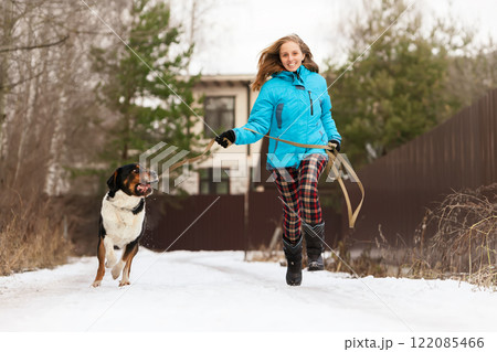 Smiling woman running with her sennenhund dog on snowy path, enjoying a winter walk. Capturing friendship, activity, and outdoor fun in cold and cheerful environment. 122085466
