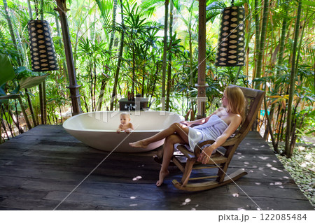 Happy baby boy have fun in bathtub, young mother relax on rocking chair in outside bathroom on veranda with beautiful tropical garden view at luxury villa on summer family vacation. Happy baby boy have fun in bathtub, young mother relax on rocking chair in outside bathroom on veranda with beautiful tropical garden view at luxury villa on summer family vacation. 122085484