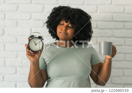 Happy african american woman holding alarm clock amazed with open mouth and surprise happy face at home, on brick background. Being late and morning time and time passing quickly concept 122090530
