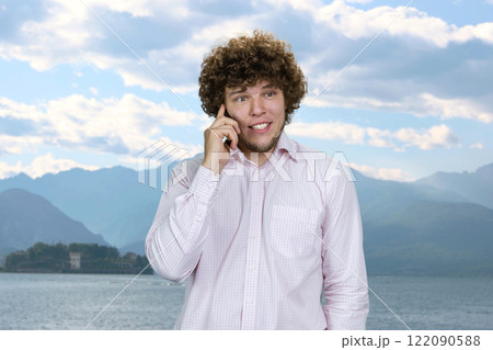 Happy smiling caucasian man in white shirt talking on the phone on vacation. Blue nature landscape in the background. Sea and mountains. 122090588