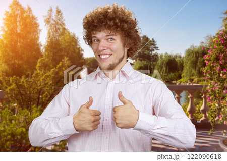 Happy smiling young man with curly hair shows both thumbs up. Sunny summer park in the background. Happy smiling young man with curly hair shows both thumbs up. Sunny summer park in the background. 122090618