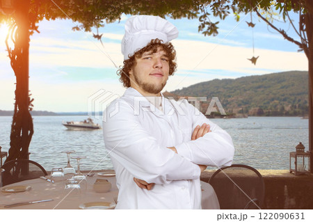 Portrait of a young male cook with folded arms standing on the beach. Evening summer sea landscape in the background. 122090631