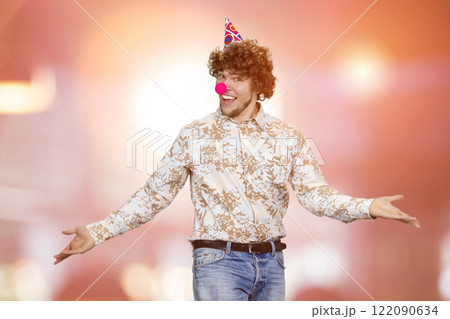 Portrait of happy young guy with clown nose and cone on his head spread his arms. Red glowing lights in the background. Portrait of happy young guy with clown nose and cone on his head spread his arms. Red glowing lights in the background. 122090634