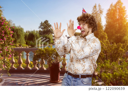 Happy young clown with curly hair wearing a red nose fooling around outdoors. Summer park in the background. 122090639
