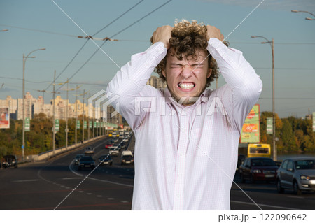 Portrait of a young man with curly hair having nervous breakdown outdoors. City street in the background. Portrait of a young man with curly hair having nervous breakdown outdoors. City street in the background. 122090642