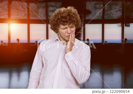 Young man with curly hair touching his mouth with hand because of toothache. Indoor background with evening sunset. Young man with curly hair touching his mouth with hand because of toothache. Indoor background with evening sunset. 122090654