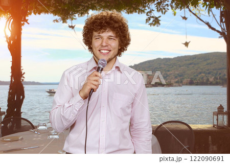 Handsome young man with curly hair singing song using microphone outdoors on vacation. Evening sea beach in the background. Handsome young man with curly hair singing song using microphone outdoors on vacation. Evening sea beach in the background. 122090691