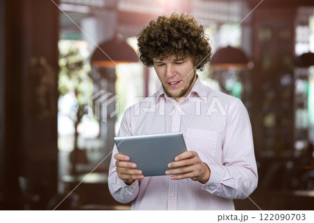 Portrait of a young man with curly hair playing a video game on the tablet pc device. Indoor restaurant in the background. Portrait of a young man with curly hair playing a video game on the tablet pc device. Indoor restaurant in the background. 122090703