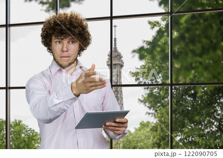 Portrait of a young caucasian worker with curly hair holding tablet pc device pointing at something. Checkered window with tv tower in the background. 122090705